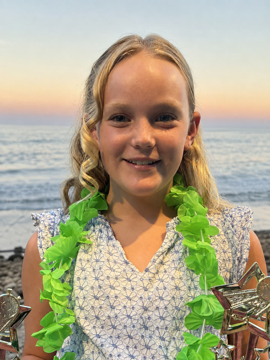 Malia smiling at the beach at sunset, wearing a bright green flower lei and holding two gold star trophies, with the ocean behind her.